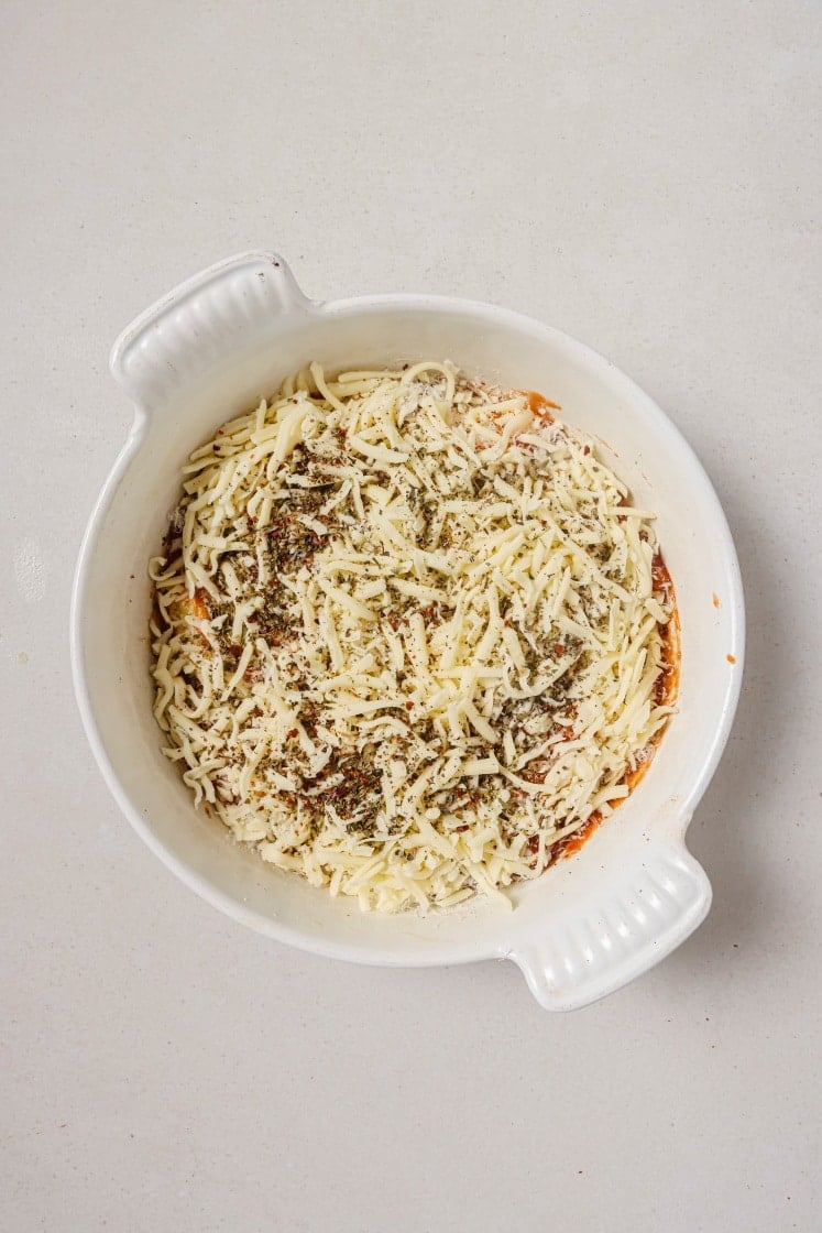 A round, white dish filled with shredded cheese topped with herbs and seasoning. The dish is set against a light gray background.