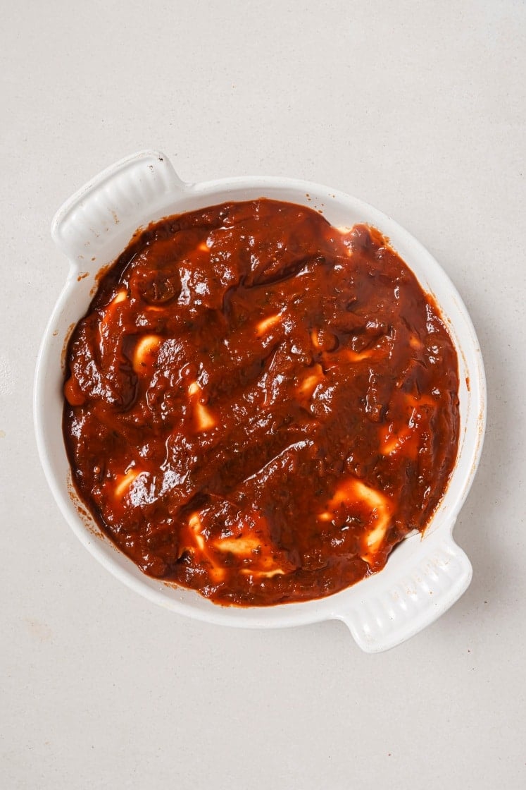 White dish filled with dark red sauce with cheese melted on top, creating a marbled effect. The dish is on a plain, light-colored surface.