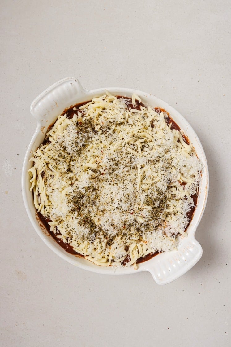 A round, white baking dish filled with lasagna, topped with melted cheese, herbs, and sauce, sits on a light countertop. The dish has two handles on opposite sides.