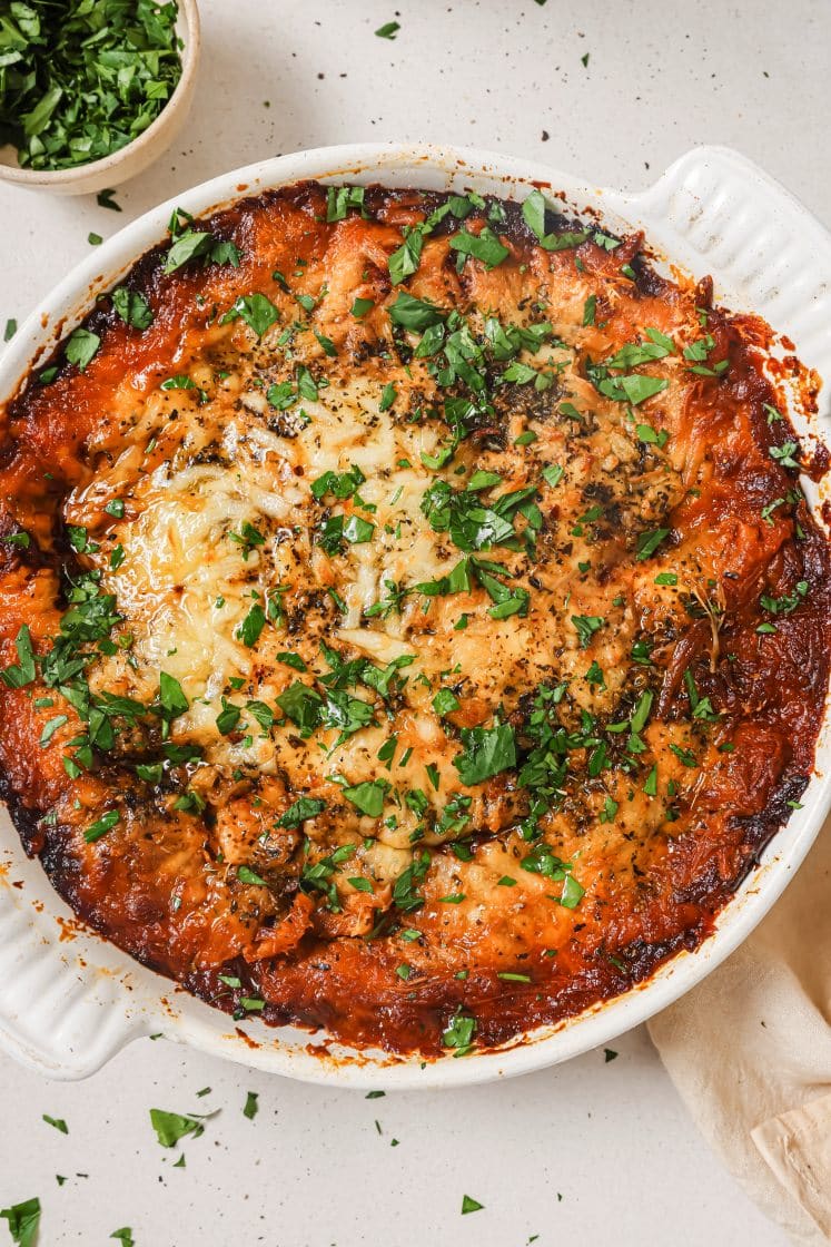 A baked dish in a round white casserole dish, topped with browned cheese and garnished with chopped parsley. The edges are crispy and slightly charred. A small bowl of chopped parsley is next to the dish on the table.