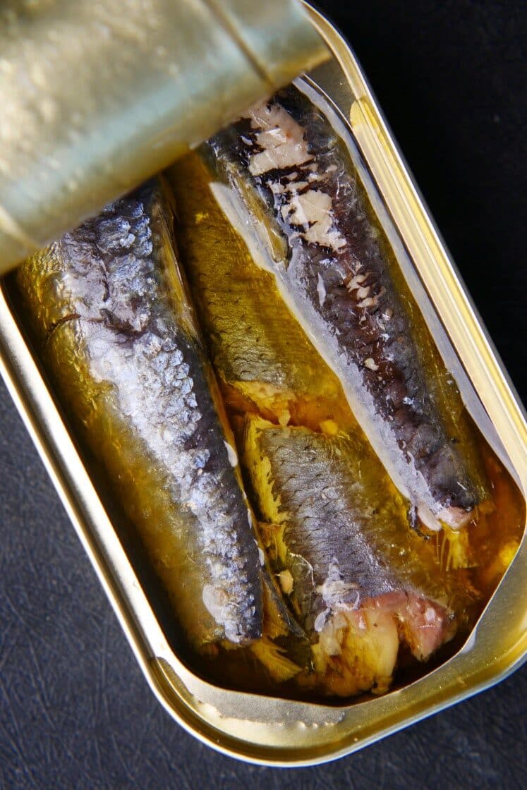 An opened tin can revealing three sardines in oil, displayed against a black background. The silver fish are neatly packed, with visible textures of the skin and oil glistening around them.