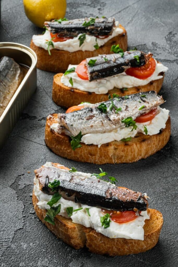 Slices of toasted bread topped with soft cheese, tomato slices, sardines, and chopped herbs are arranged in a line. A lemon sits nearby, and a tin of sardines is partially visible on the left side. The surface is gray and textured.