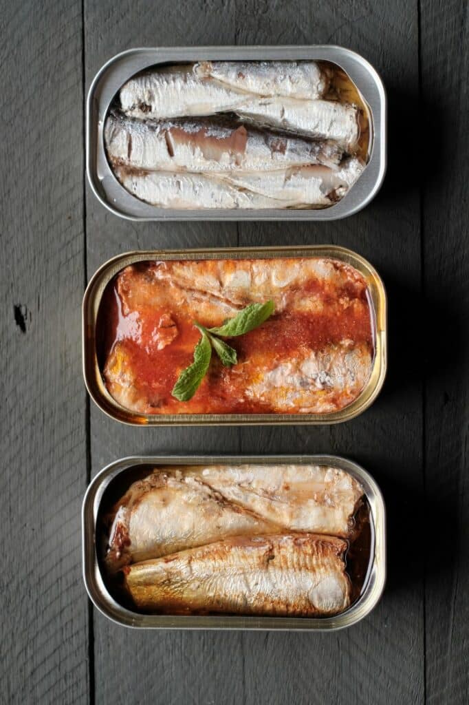 Three open tins of sardines are arranged on a dark wooden surface. The top tin contains sardines in oil, the middle tin has sardines in tomato sauce with a basil garnish, and the bottom tin has sardines in a light sauce.