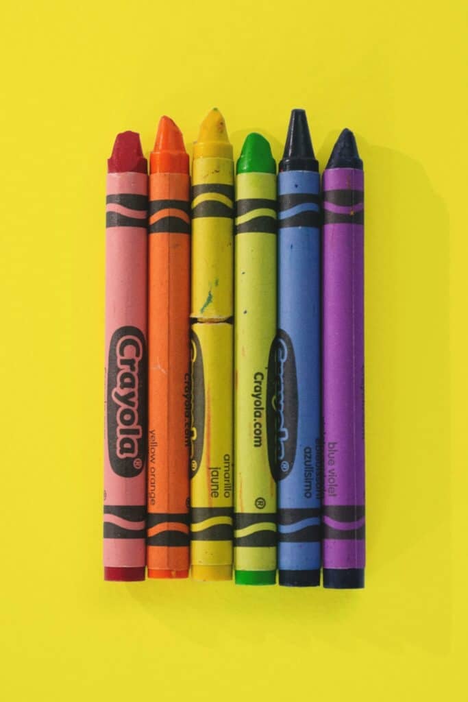 Six Crayola crayons—red, orange, yellow, green, blue, and purple—are lined up vertically on a bright yellow background.