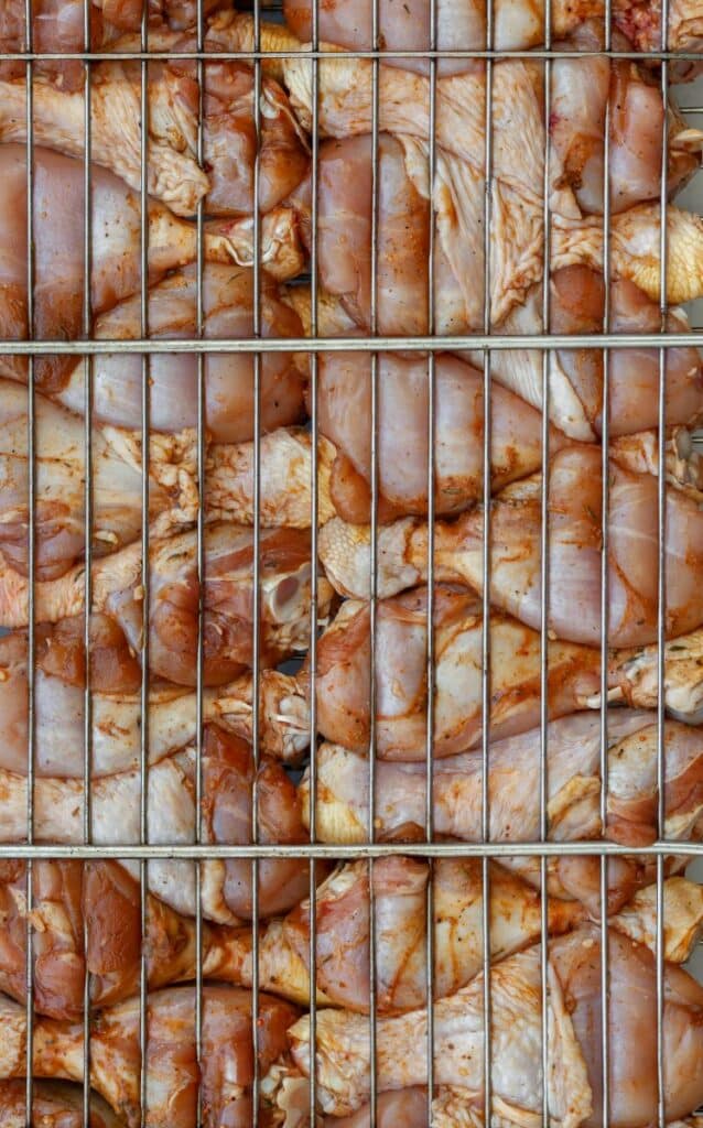 Close-up of raw, seasoned chicken pieces stacked behind a metal grill or rack, preparing for grilling or cooking. The chicken is marinated and arranged tightly within the grid.