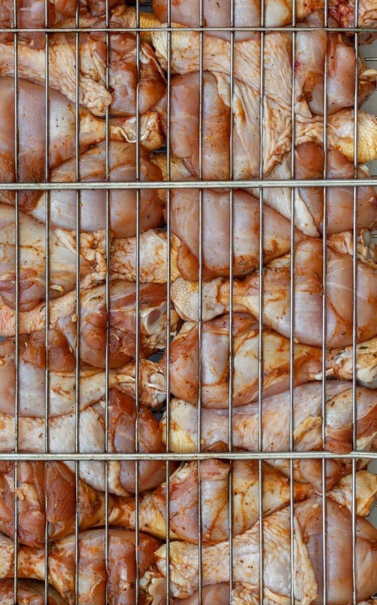 Close-up of raw, seasoned chicken pieces stacked behind a metal grill or rack, preparing for grilling or cooking. The chicken is marinated and arranged tightly within the grid.