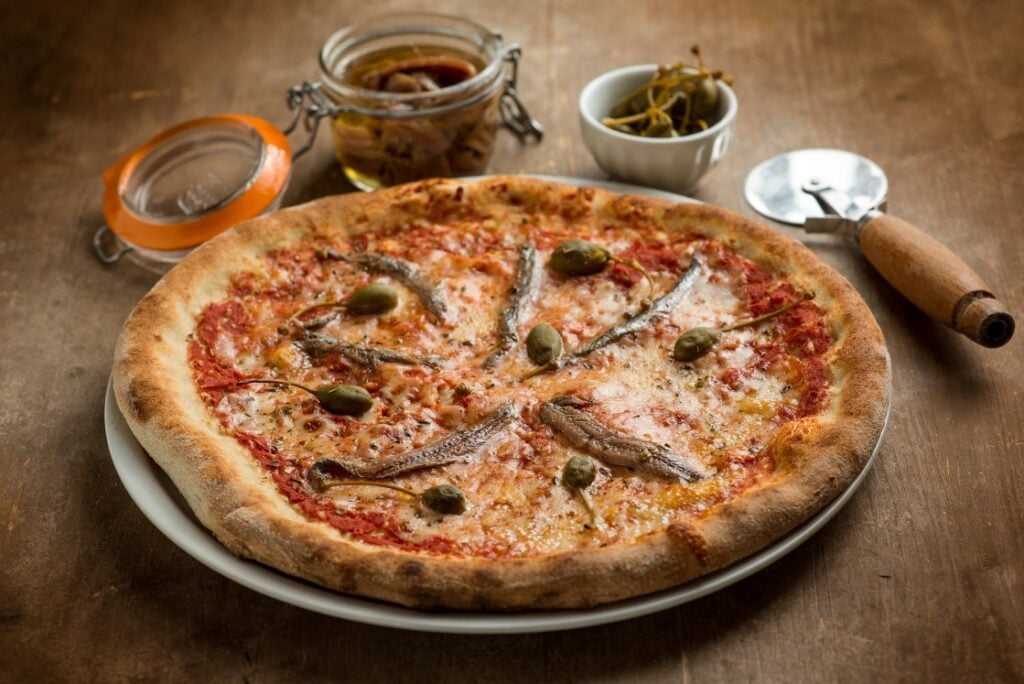 A baked pizza topped with anchovies, capers, and cheese sits on a plate. Behind it are a jar of anchovies, a small bowl of capers, and a pizza cutter on a wooden table.