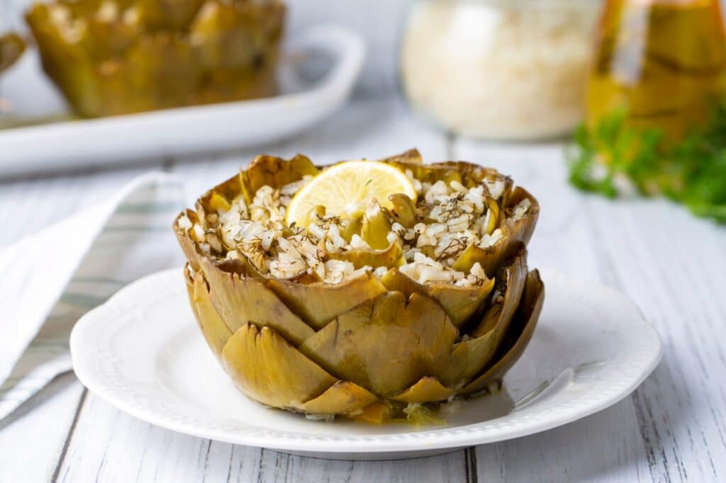 A cooked artichoke stuffed with rice and herbs, garnished with a lemon slice, sits on a white plate. A fork rests beside the plate on a napkin, and jars are blurred in the background on a white wooden table.