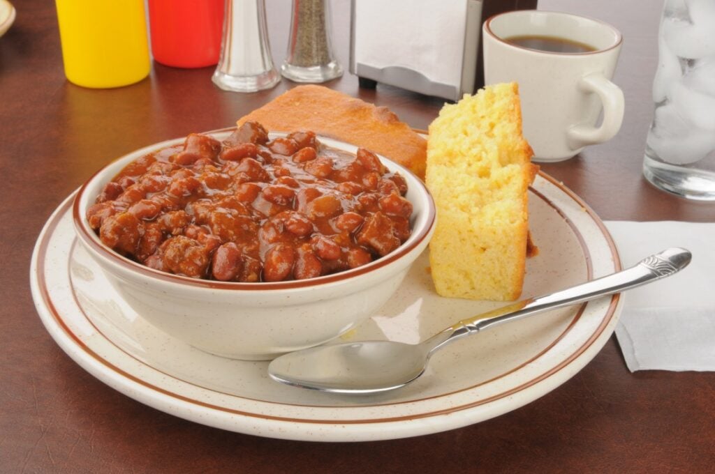 A bowl of chili with beans sits on a plate next to two slices of cornbread and a spoon. In the background are a cup of coffee, a glass of ice water, and condiments on a brown table.