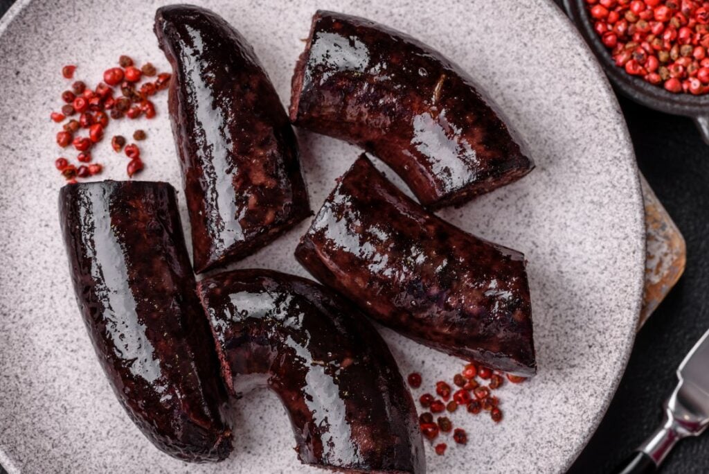 Five pieces of dark brown blood sausage are arranged on a light gray plate, with scattered pink peppercorns beside them. The sausages have a glossy, rich appearance.