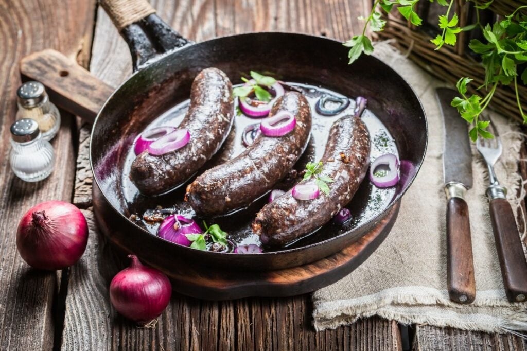 A rustic skillet with four cooked blood sausages garnished with sliced red onions and herbs, sitting on a wooden table with whole red onions, cutlery, salt, pepper, and fresh parsley nearby.