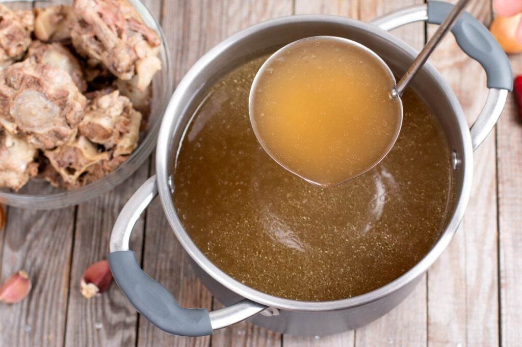 A pot of golden brown broth with a ladle lifting some liquid, next to a bowl filled with cooked bones, on a wooden surface.