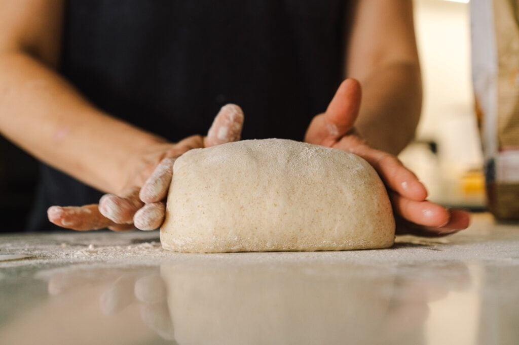 Person shaping a ball of dough on a floured surface with both hands. The background is out of focus, drawing attention to the dough and hands in the foreground.