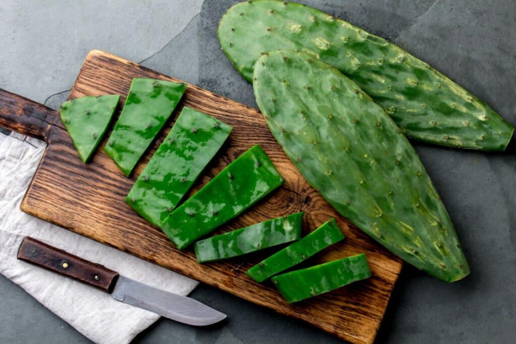 Two whole cactus pads and several sliced pieces of cactus are arranged on a wooden cutting board next to a knife, all set on a gray stone surface.