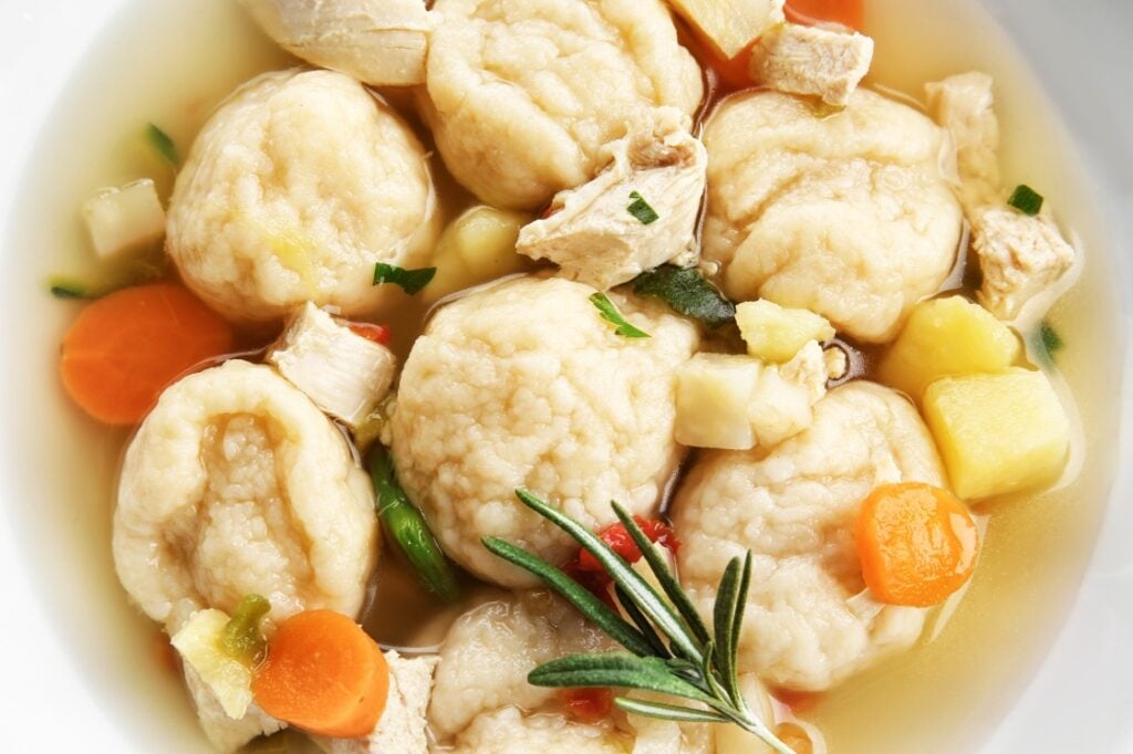A close-up of a bowl of matzo ball soup with fluffy matzo balls, diced carrots, potatoes, herbs, and pieces of chicken in clear broth, garnished with a sprig of rosemary.