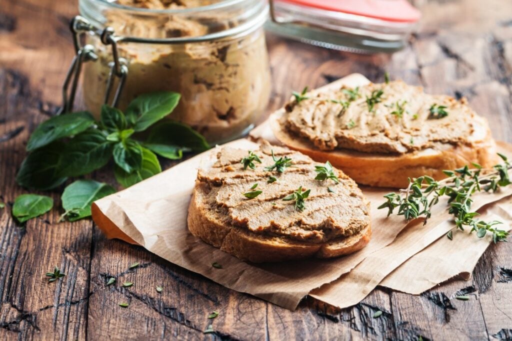 Two slices of bread topped with pâté and fresh herbs rest on brown parchment paper. A jar of pâté and a sprig of fresh basil are in the background on a rustic wooden table.
