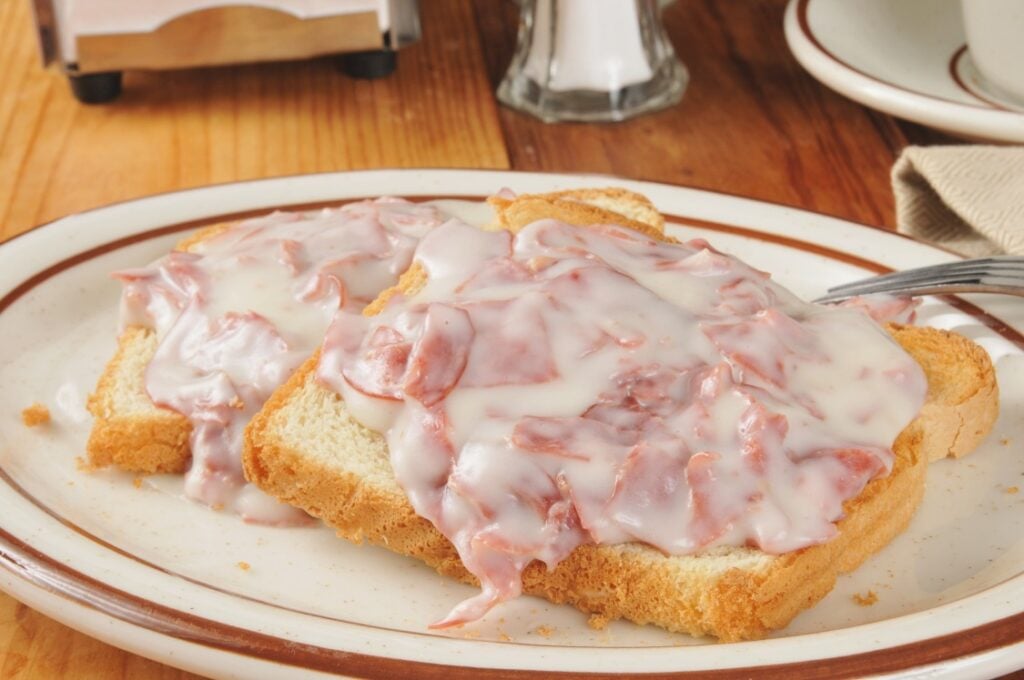 Two slices of toasted bread covered with creamy, chipped beef in white gravy, served on a white plate with a brown rim, on a wooden table.