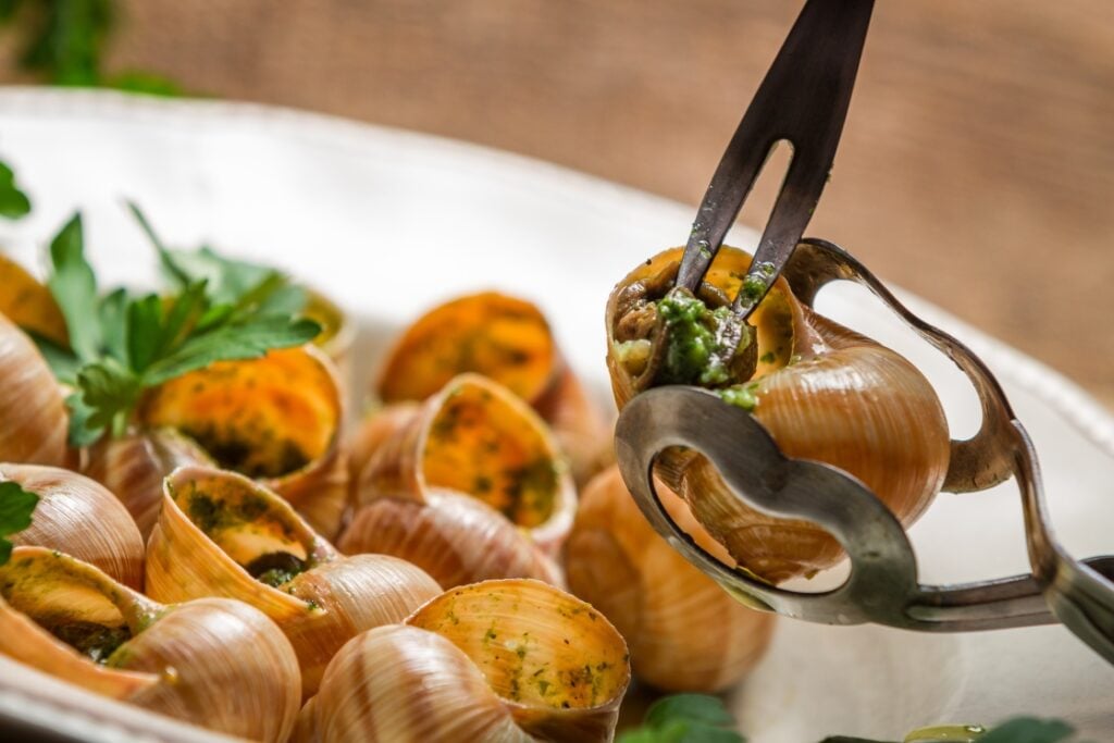 A close-up of escargot shells filled with herb butter, served in a white dish. A fork and tongs are holding one escargot, and fresh parsley is used as garnish.