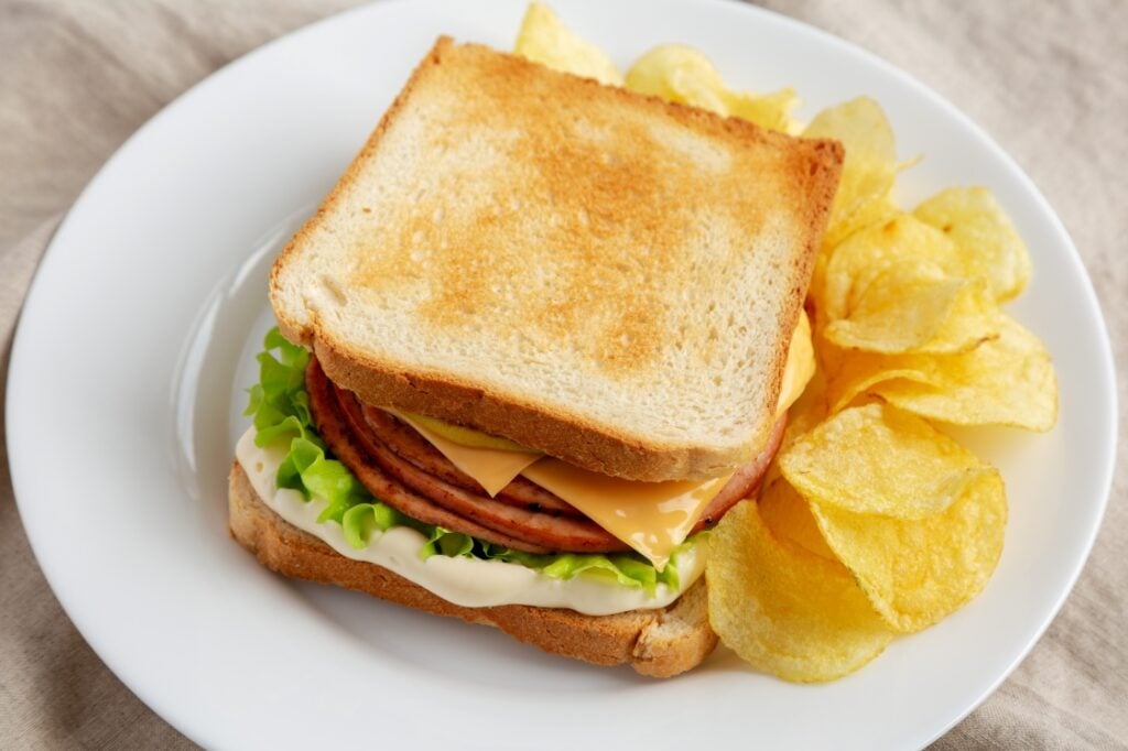 A sandwich with toasted bread, lettuce, cheese, mayonnaise, and sliced meat is served on a white plate with a side of potato chips.