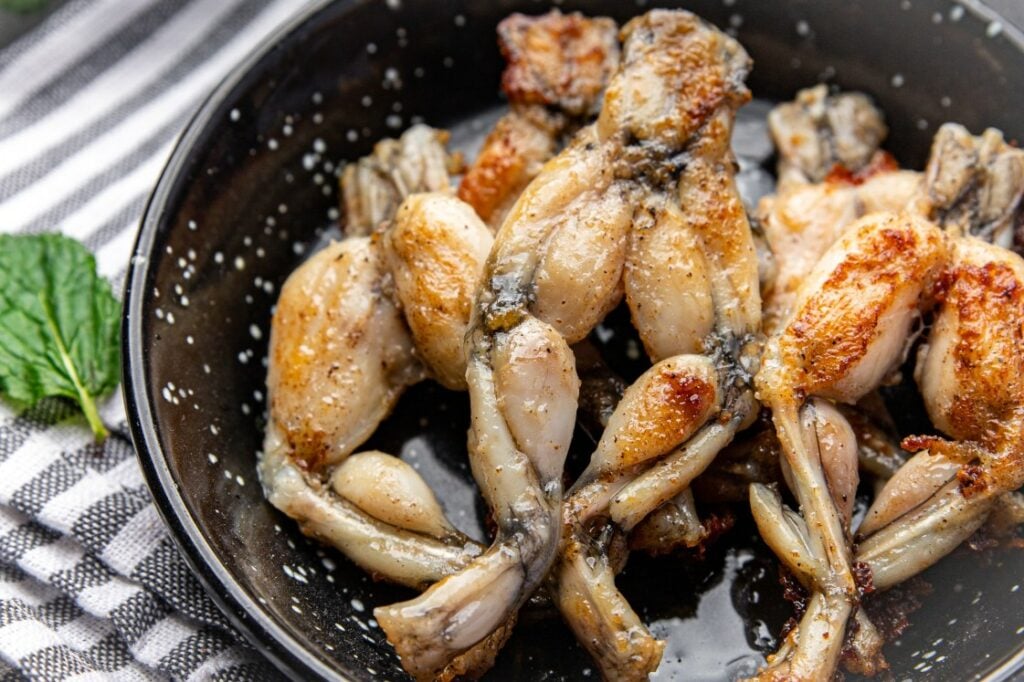 A black bowl filled with seasoned and cooked frog legs, lightly browned and garnished with coarse salt, sits on a striped cloth next to a green mint leaf.