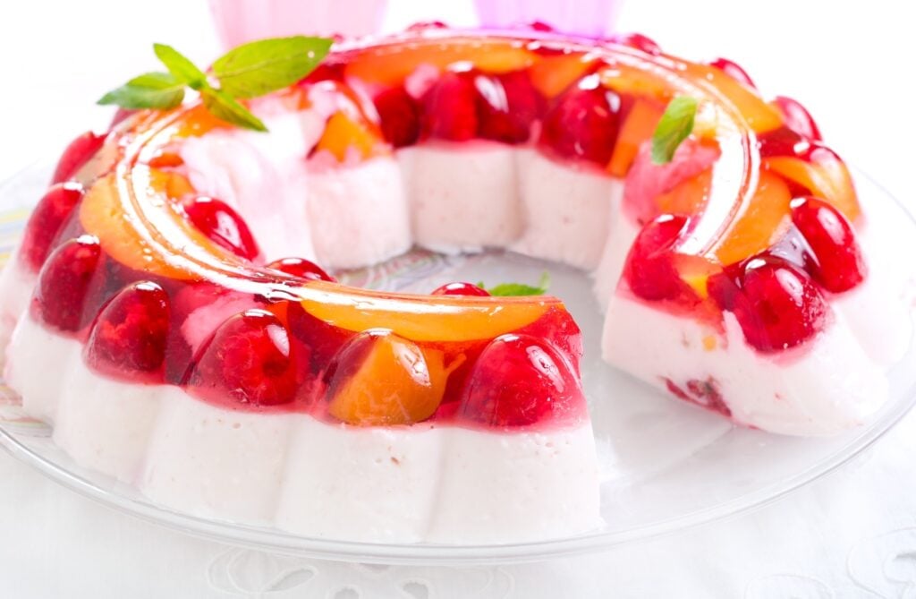 A gelatin dessert ring with layers of pink jelly, strawberries, and peach slices, garnished with mint leaves, is displayed on a clear glass plate. A slice has been removed to show the layered texture.