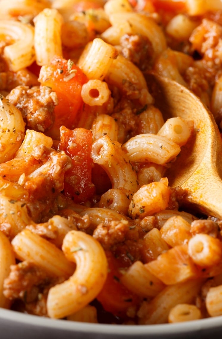 Close-up of cooked elbow macaroni mixed with ground beef, diced tomatoes, and sauce, being stirred with a wooden spoon. The dish appears hearty and well-seasoned.