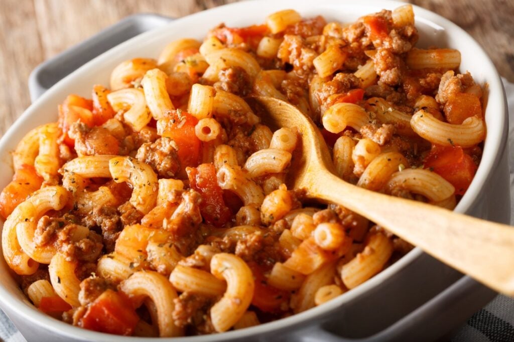A close-up of a casserole dish filled with elbow macaroni, ground meat, tomato sauce, and diced tomatoes, with a wooden spoon resting inside.