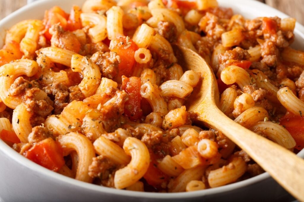 A close-up of American goulash with elbow macaroni, ground beef, and diced tomatoes in a white dish. A wooden spoon rests in the pasta, mixing the ingredients together.