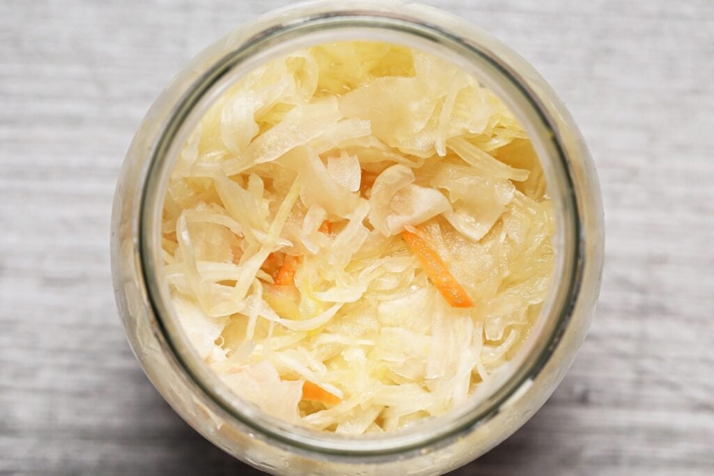 Top-down view of a glass jar filled with shredded sauerkraut, including pale cabbage and small orange carrot strips, on a light gray surface.