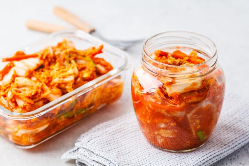 A glass jar and a rectangular container filled with homemade kimchi, featuring napa cabbage and carrots in a spicy red seasoning, sit on a folded gray cloth with wooden-handled utensils in the background.