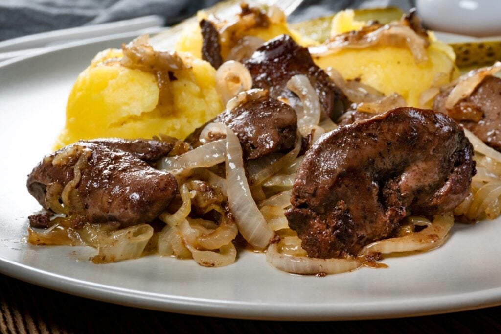 A plate with cooked liver and sautéed onions, served alongside scoops of mashed potatoes. The food looks freshly prepared and garnished, with a savory, home-cooked appearance.