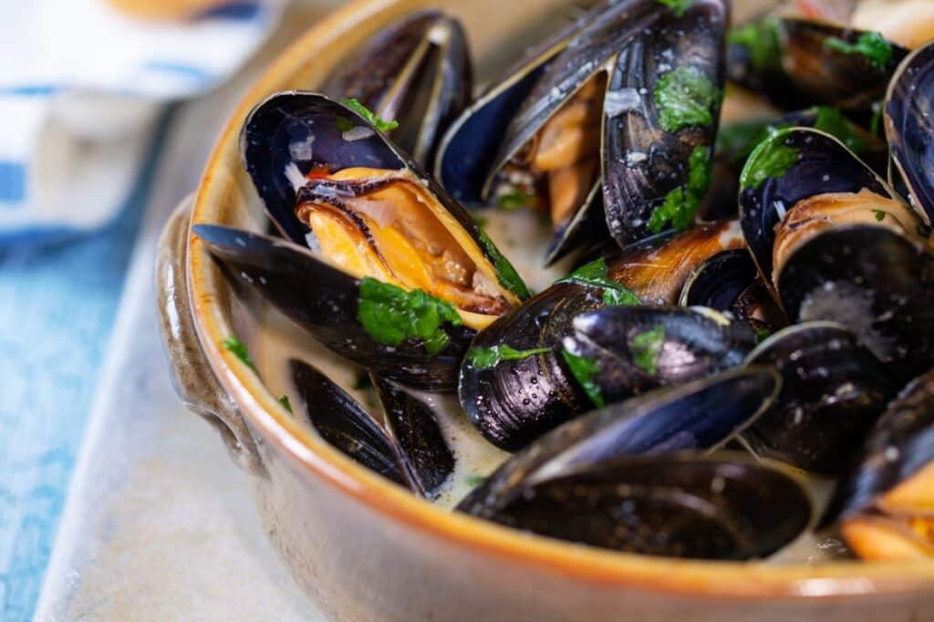 A bowl of steamed mussels in broth, garnished with chopped parsley. The mussels' dark shells are open, revealing the orange flesh inside. The dish is served in a rustic brown ceramic bowl.