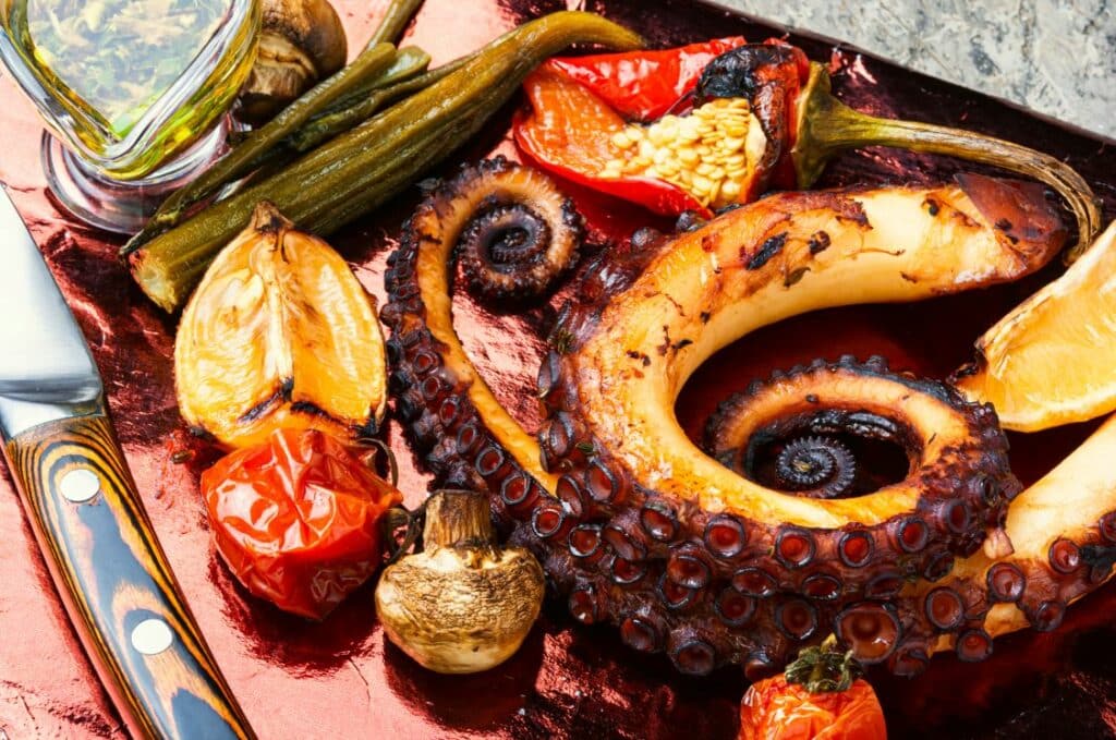 Grilled octopus tentacle served with roasted tomatoes, mushrooms, okra, red pepper, and a lemon wedge on a metallic surface. A knife and a small glass bowl of green sauce are also visible.