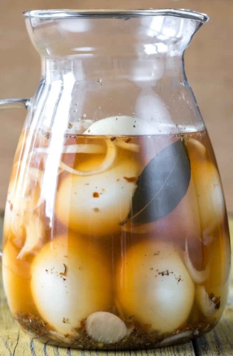 A glass pitcher filled with whole hard-boiled eggs, onion slices, bay leaf, and spices submerged in brine, sitting on a wooden surface.