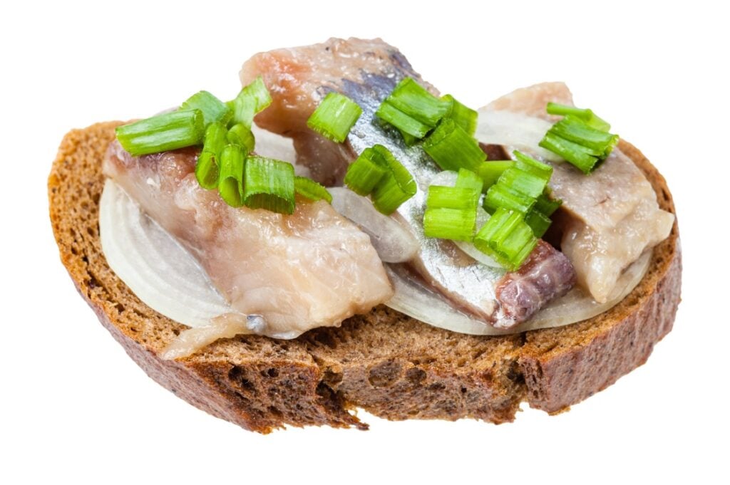 A slice of dark rye bread topped with pieces of pickled herring, sliced white onion, and chopped green onions, isolated on a white background.