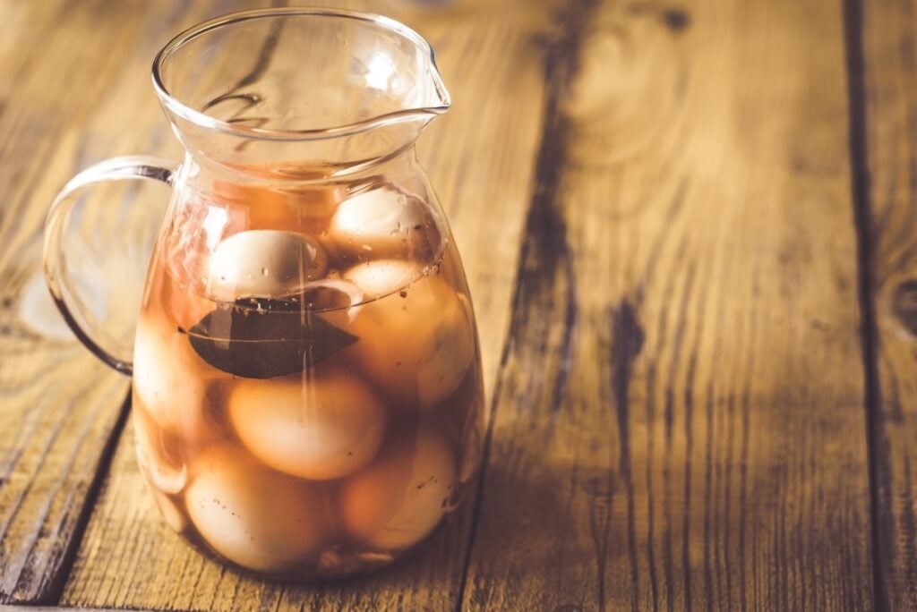 A glass pitcher filled with eggs and a brownish brine, containing a bay leaf, sits on a rustic wooden table.