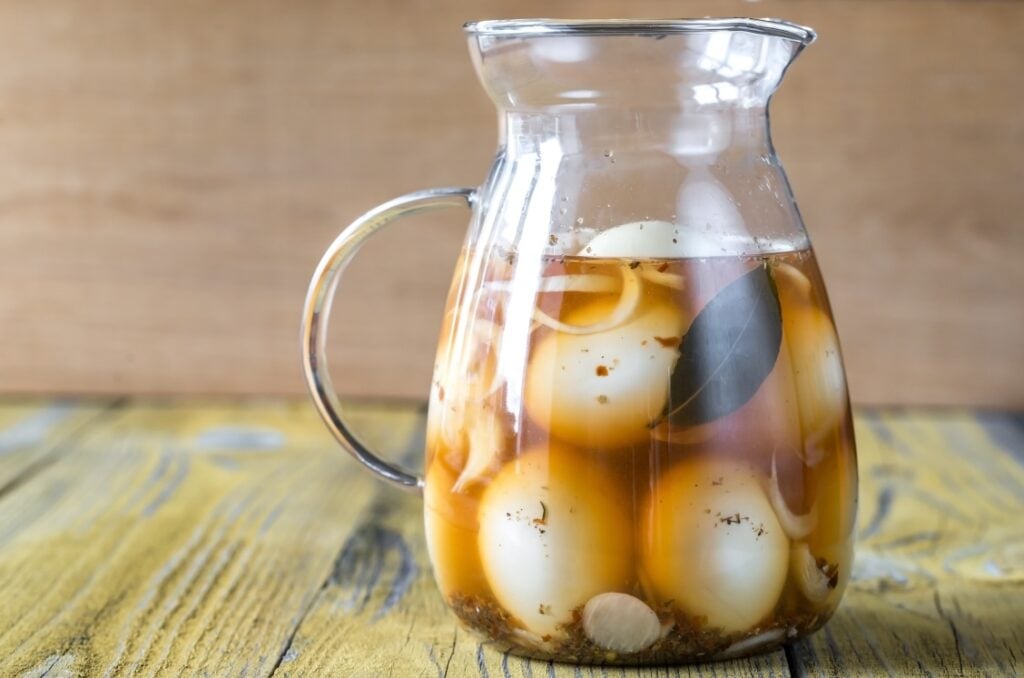 A glass pitcher filled with pickled eggs, onion slices, spices, and a bay leaf in a brine, sitting on a rustic yellow wooden surface with a wooden background.