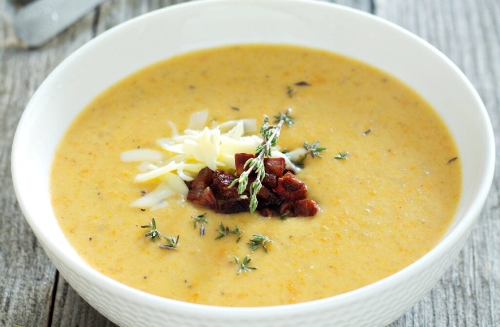 A bowl of creamy soup garnished with shredded cheese, crispy bacon pieces, and fresh thyme, served in a white bowl on a rustic wooden table.