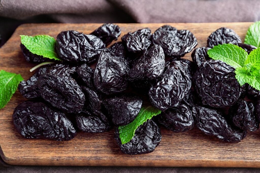 A pile of dried prunes arranged on a wooden cutting board, garnished with fresh mint leaves, against a brown fabric background.
