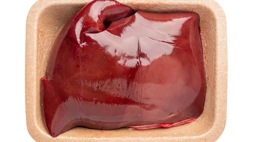 A raw beef liver—one of those foods people are afraid to cook—rests in a beige, rectangular tray, its glossy, reddish-brown surface glistening under the light.