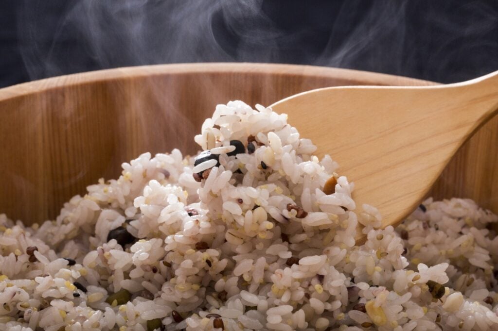 A close-up of steaming mixed rice with grains and beans, being scooped with a wooden spoon from a wooden bowl.
