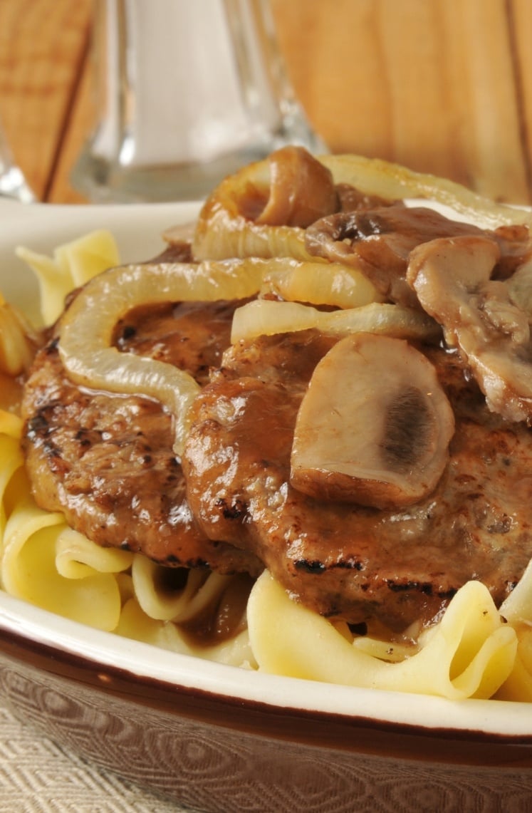 A plate of Salisbury steak topped with sautéed onions and mushrooms, served over a bed of egg noodles, with a brown gravy sauce.