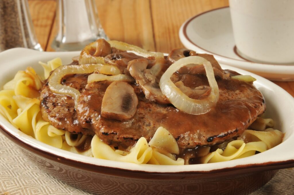 A plate of Salisbury steak topped with sautéed onions and mushrooms in brown gravy, served over egg noodles, with a cup and salt and pepper shakers in the background.