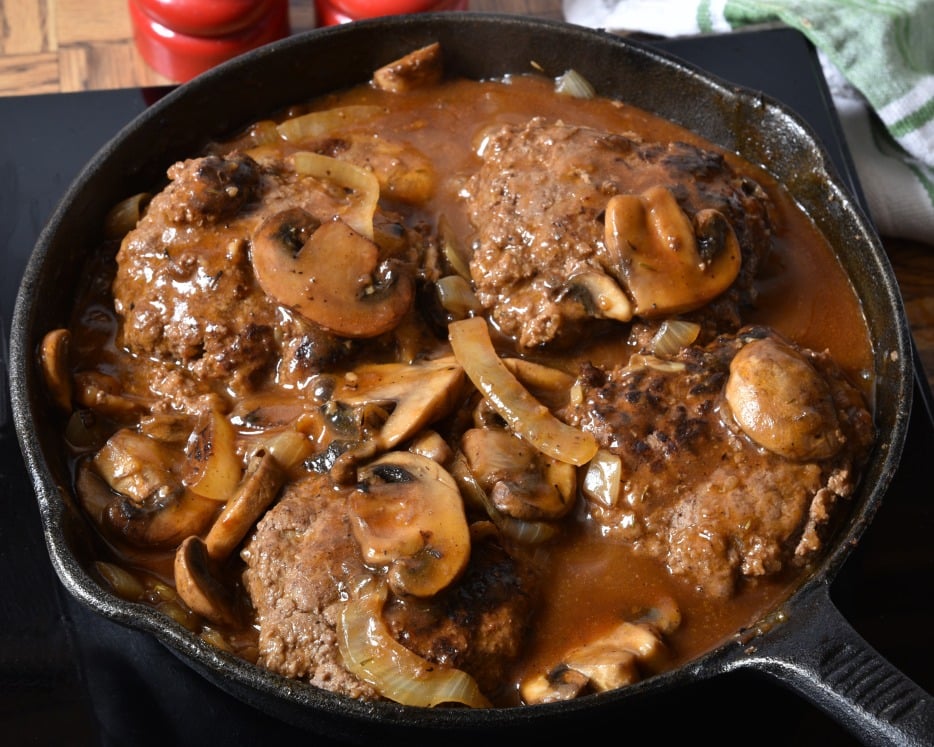 A cast-iron skillet filled with Salisbury steak patties simmering in brown gravy with sliced mushrooms and onions. The dish appears hot and freshly cooked, with rich sauce covering the meat and vegetables.