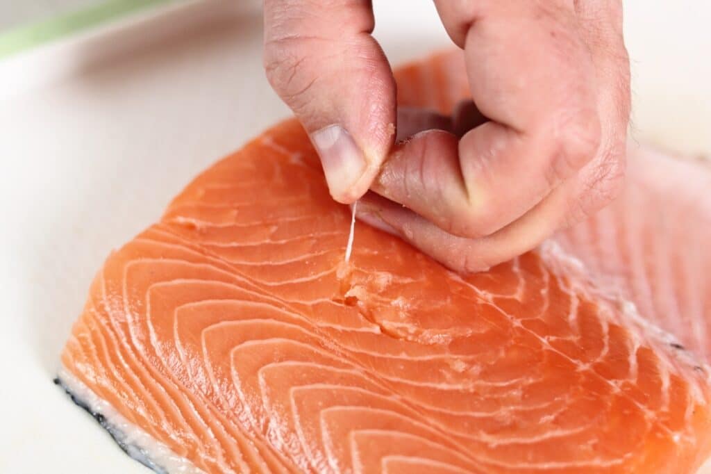 A close-up of a hand pulling a small bone from a raw salmon fillet on a white surface.