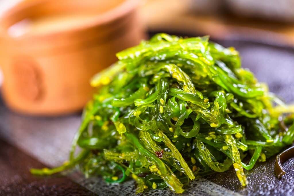 A close-up of a bright green seaweed salad garnished with sesame seeds, served on a plate with a blurred bowl in the background.