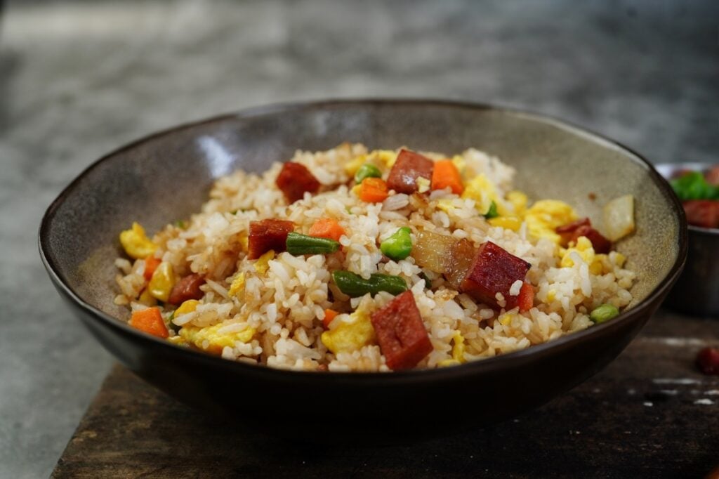 A bowl of fried rice with diced vegetables, scrambled eggs, and chunks of meat, served in a brown ceramic bowl on a rustic surface.