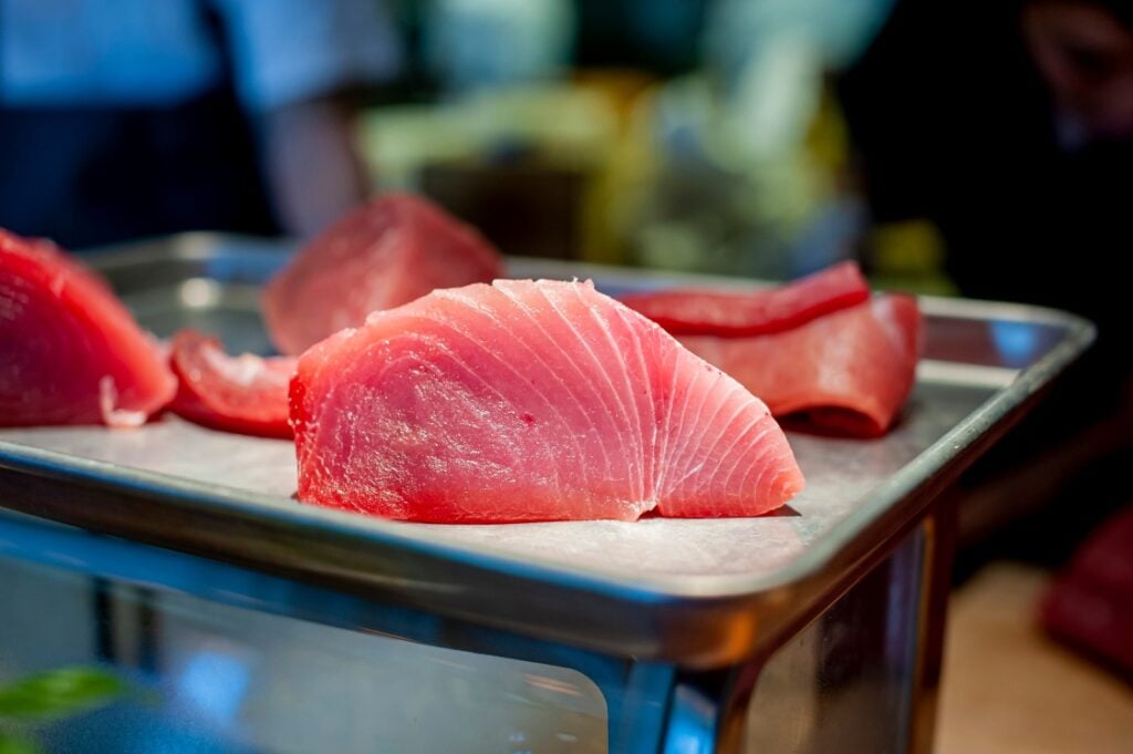 A close-up of fresh, raw tuna sashimi pieces displayed on a metal tray, with a blurred background in what appears to be a seafood market or sushi restaurant.