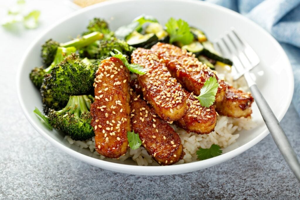 A bowl of sesame-crusted tempeh strips on white rice, served with roasted broccoli and zucchini, garnished with fresh cilantro. A fork rests in the bowl.