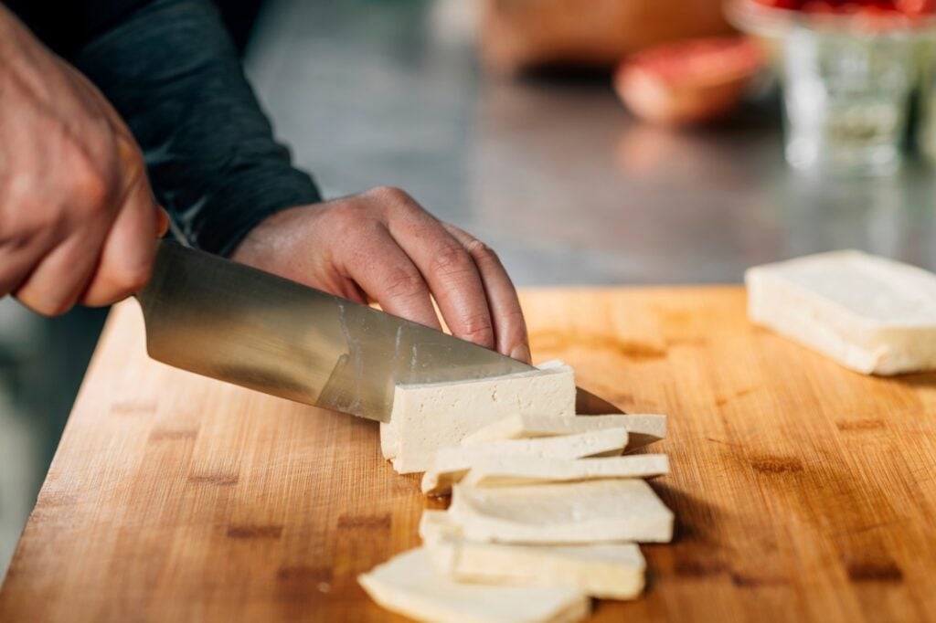 A person slices a block of tofu with a large kitchen knife on a wooden cutting board.
